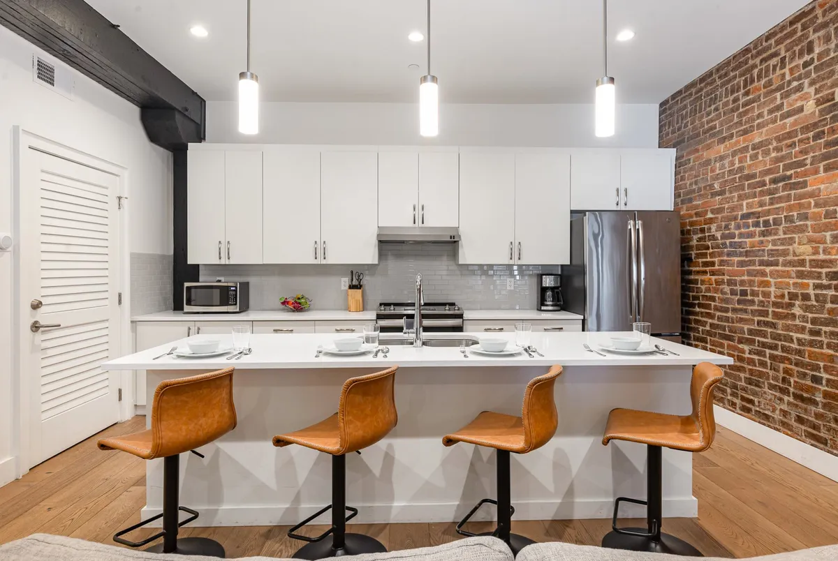 A contemporary kitchen scene featuring a spacious kitchen island with four bar stools, exposed brick wall, and gleaming stainless steel appliances. Ample counter space is visible, and the overall mood is both cozy and lively, perfect for both cooking and entertaining.
