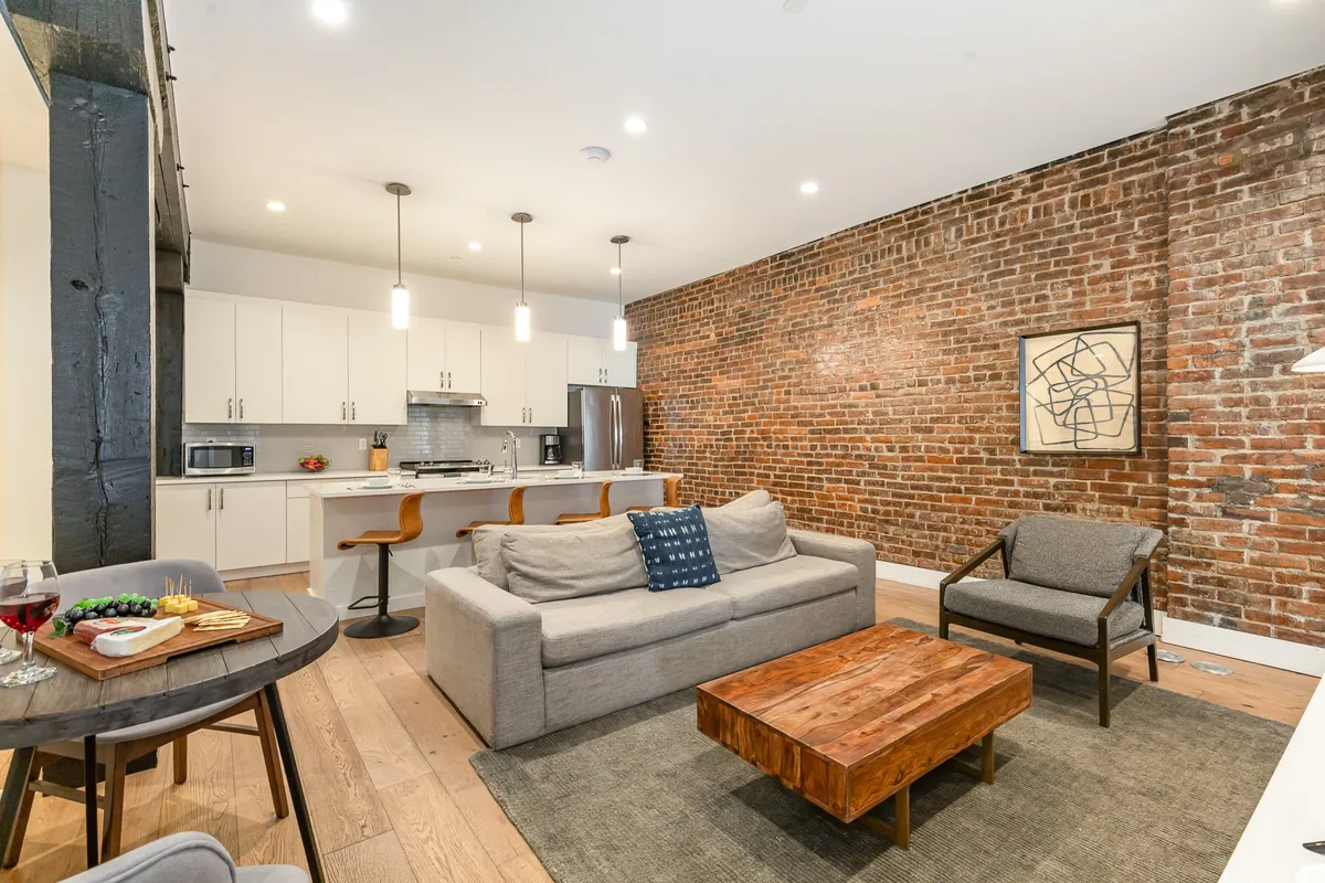 A broader view of an open living and kitchen area featuring a comfortable sofa, a wooden coffee table, exposed brick wall, and a modern kitchen in the background. The setting feels warm, inviting, and perfect for group socializing, showing how the space easily transitions from lounging to entertaining.