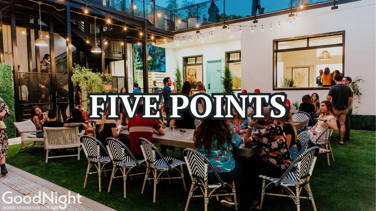 An exterior communal patio set up with a large table, surrounded by modern architecture and strung with glowing lights. Groups of people are gathered, dining and socializing, while the ambiance is lively and festive, hinting at a welcoming space for group gatherings.