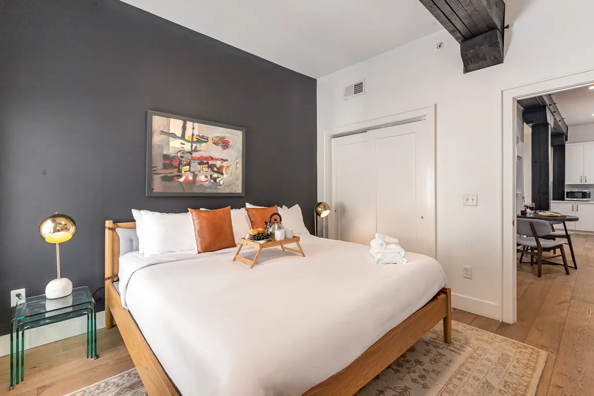 A light-filled primary bedroom featuring a king-sized bed dressed in crisp linens, with decorative artwork above the headboard. The room is styled with a bedside lamp and a tray with breakfast items, creating an inviting and restful ambiance.