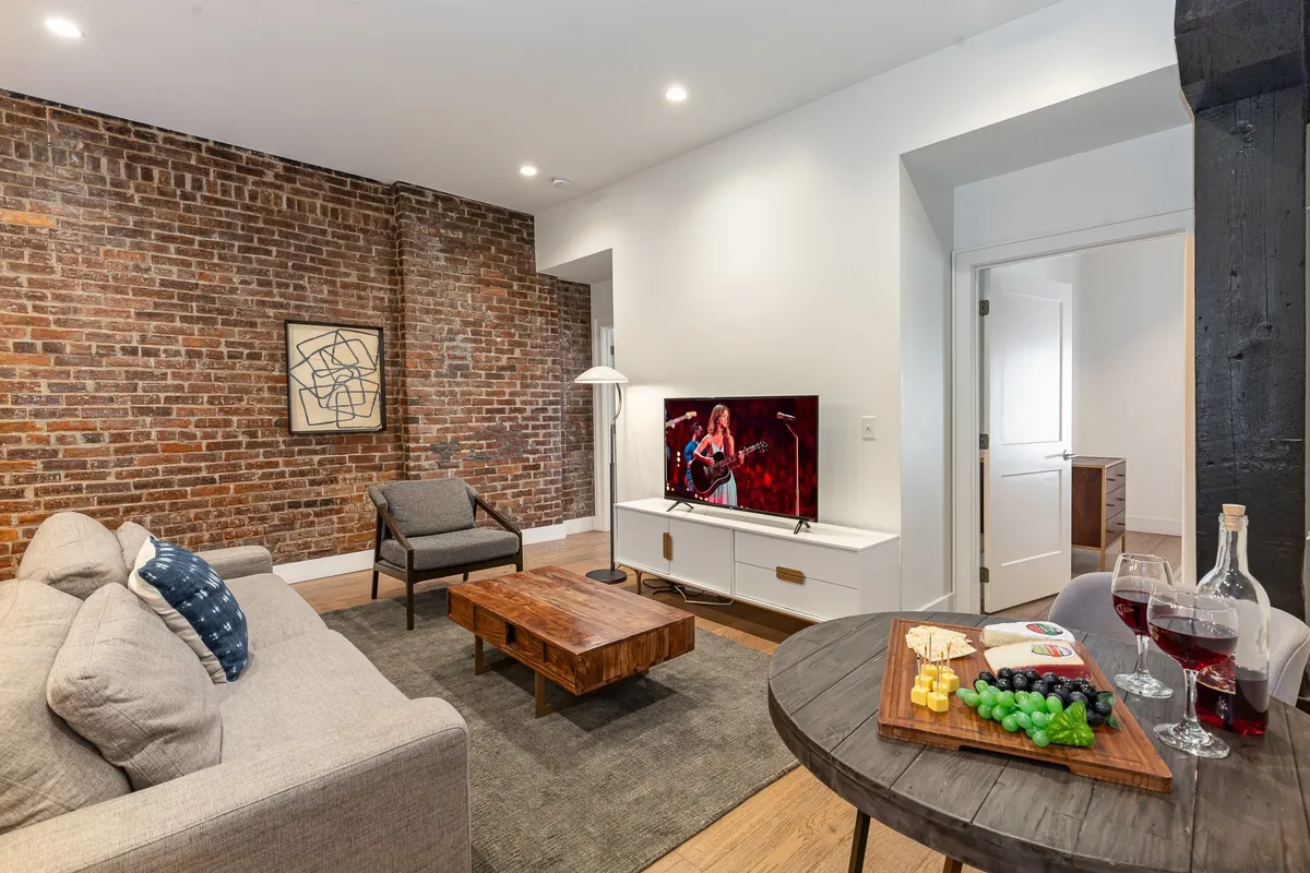 A welcoming living room with an exposed brick accent wall, a cozy sofa facing a TV that displays a performance, and a coffee table set with snacks—inviting for both social gatherings and relaxation. The ambient lighting and homey decor give the space a lively yet comfortable atmosphere.