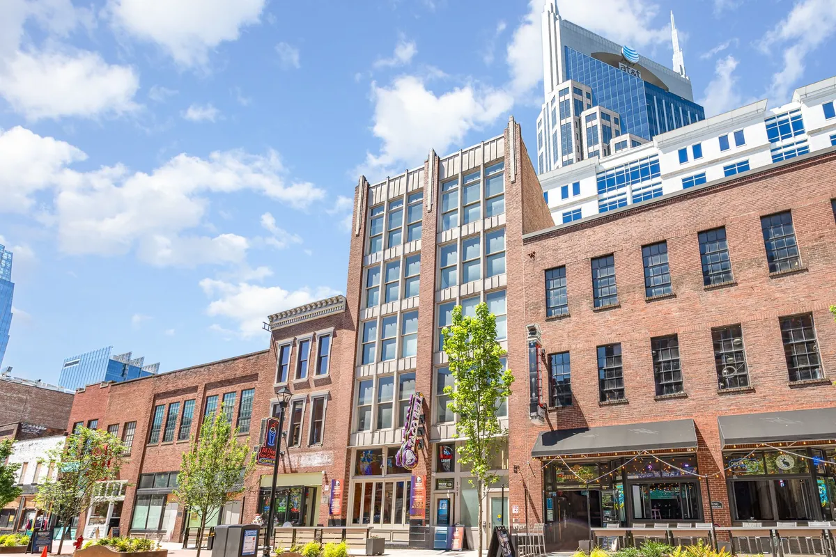 An energetic street-level shot featuring a historic brick building, modern skyscraper, outdoor seating, and groups of people dining, shopping, and socializing. The mix of old and new architecture underscores the dynamic urban atmosphere.