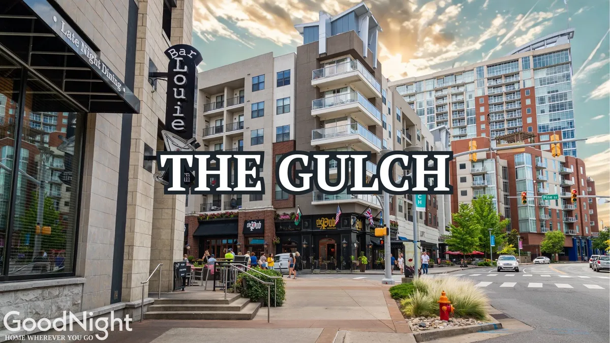A lively city street scene featuring the Bar Louie restaurant, The Gulch neighborhood signage, and modern apartments, all bustling with people dining at outdoor cafes, shopping, and socializing. The energy of the street emphasizes urban excitement and accessibility to local attractions.