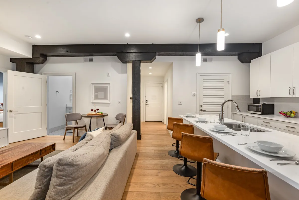 An inviting, wide-angle view that seamlessly combines the kitchen island with bar stools, a nearby dining area with a table for two, and a comfortable sofa. The modern, open-concept layout facilitates socializing and shared meals.