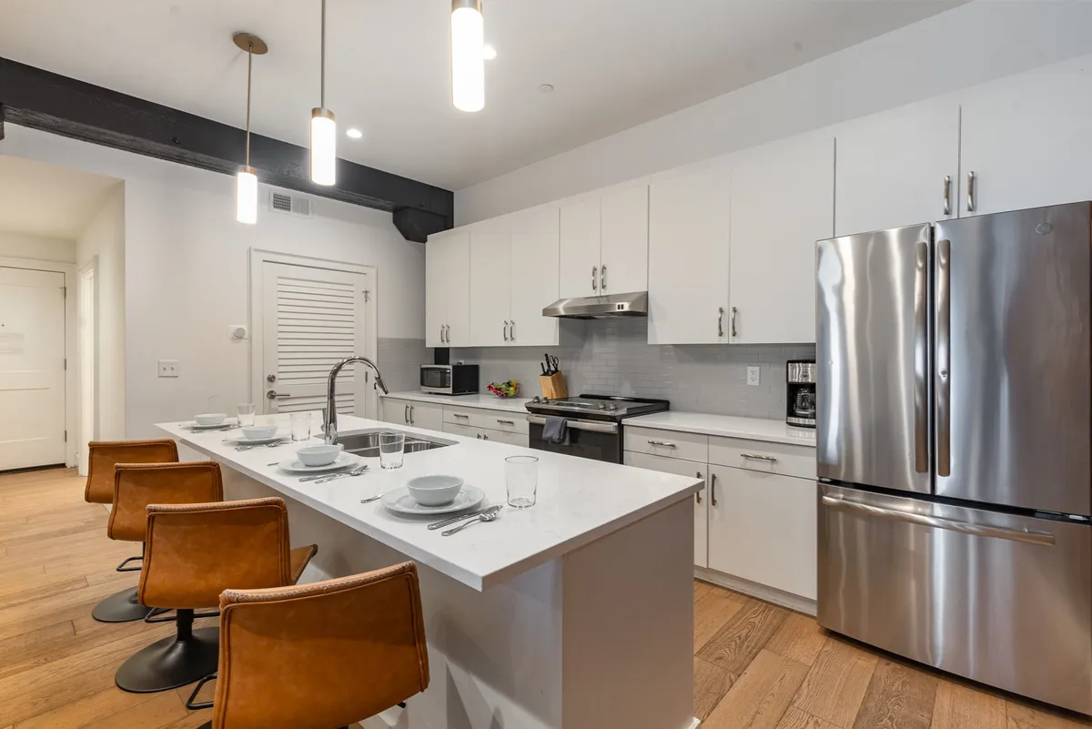 A bright, modern kitchen view with a focus on a well-appointed island, sleek bar stools, and high-end, stainless steel appliances. The setting is ready for cooking, entertaining, or casual conversation, with plenty of natural light adding to the welcoming mood.